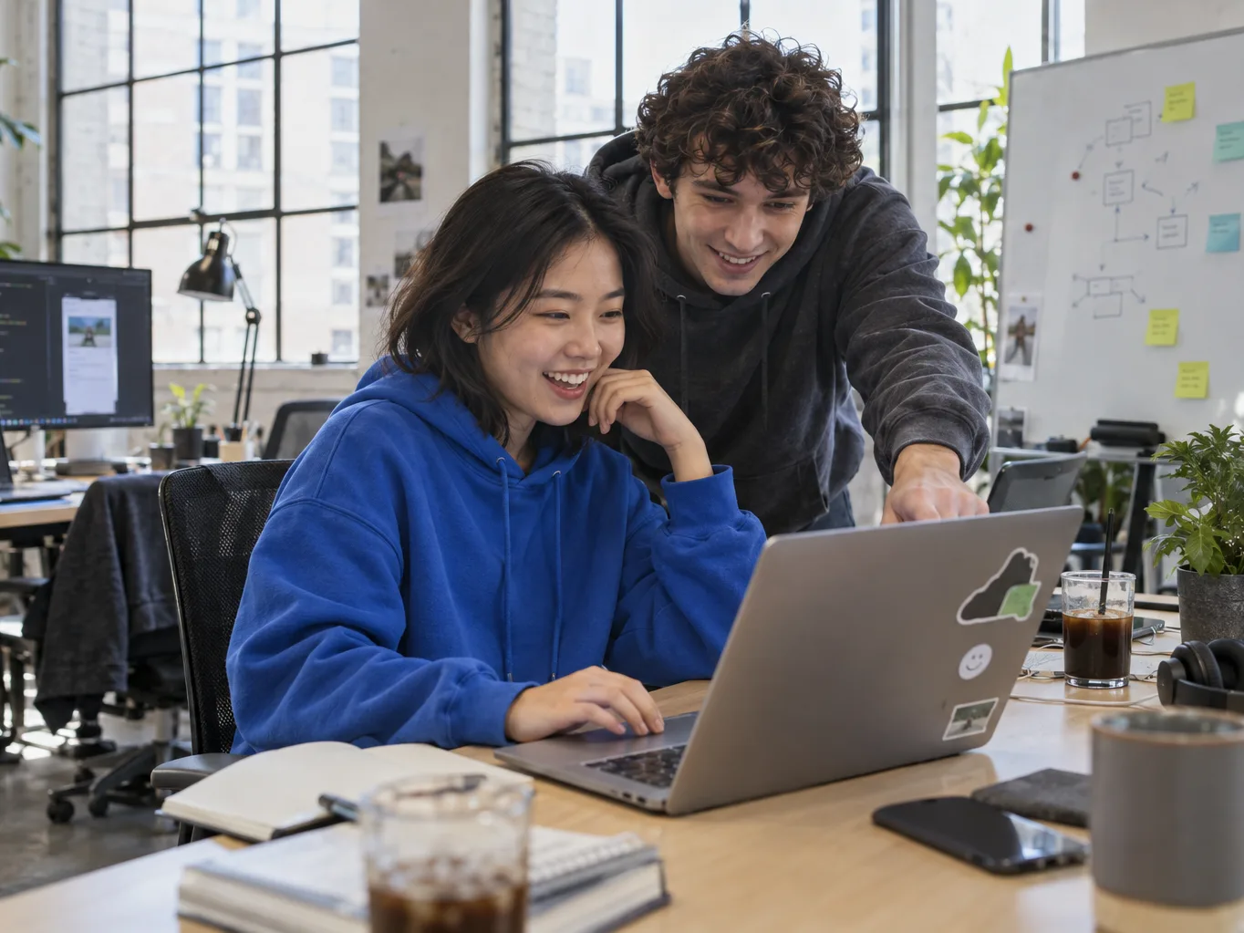 Two people share a laptop at a desk in an open office, one seated and typing while the other leans in and points at the screen. A whiteboard of diagrams stands behind them.