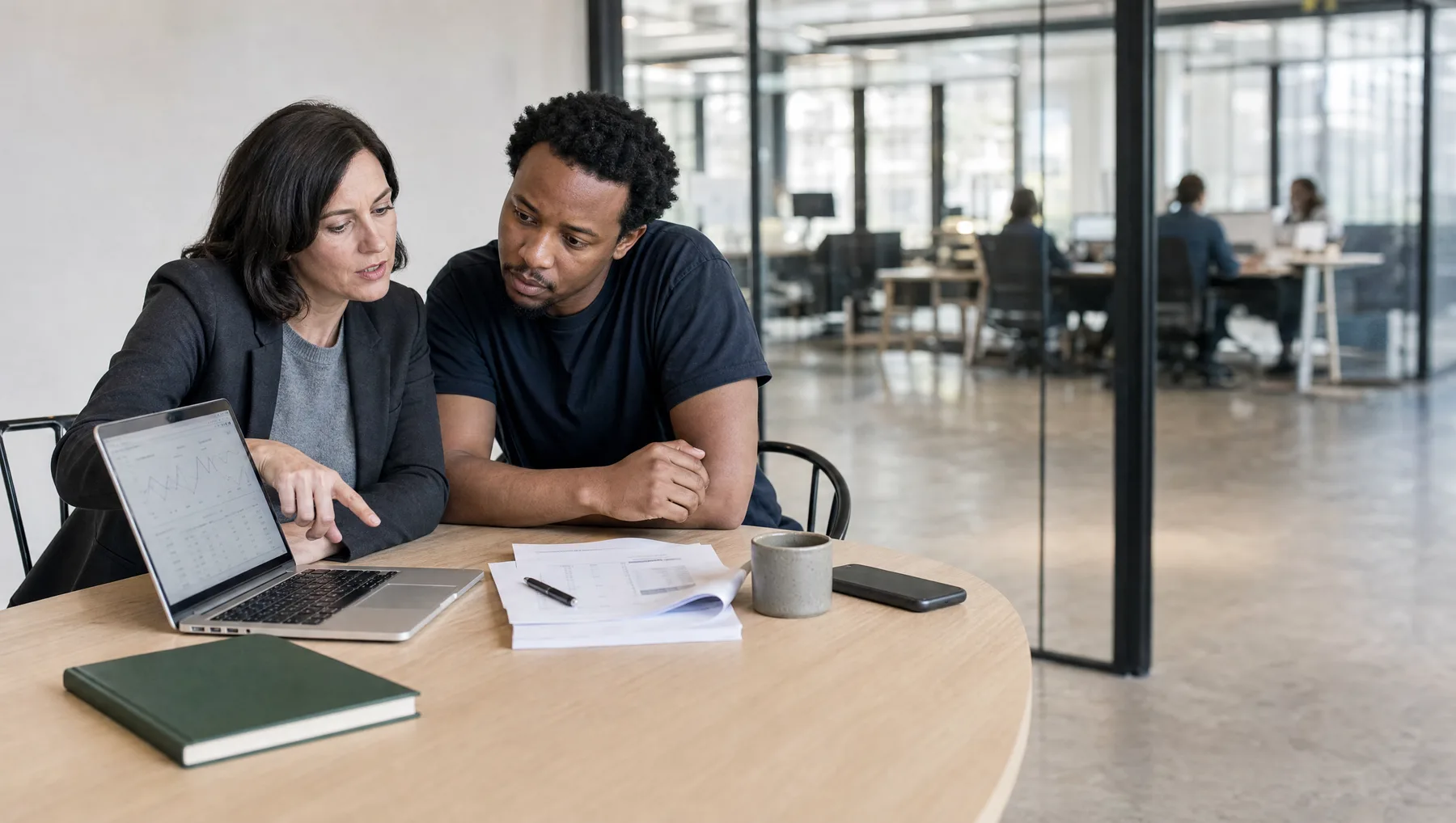 A fractional CFO in a charcoal blazer points at a line on a laptop dashboard while the founder beside her follows, printed statements and a pen on the table, a deep green notebook in the foreground and a glass-walled coworking space behind them.